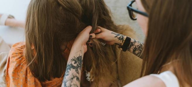 A woman with tattoos on her arm is braiding two girls' hair together.